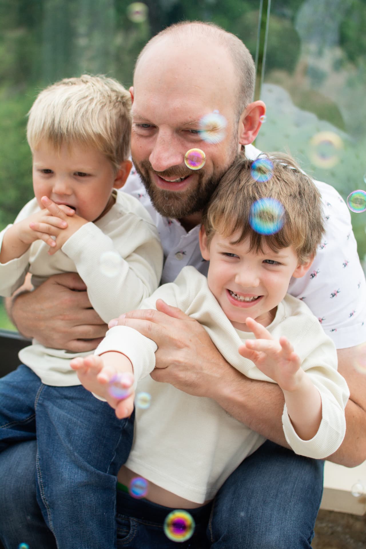 Father holding two young boys as they play with colourful bubbles on a balcony