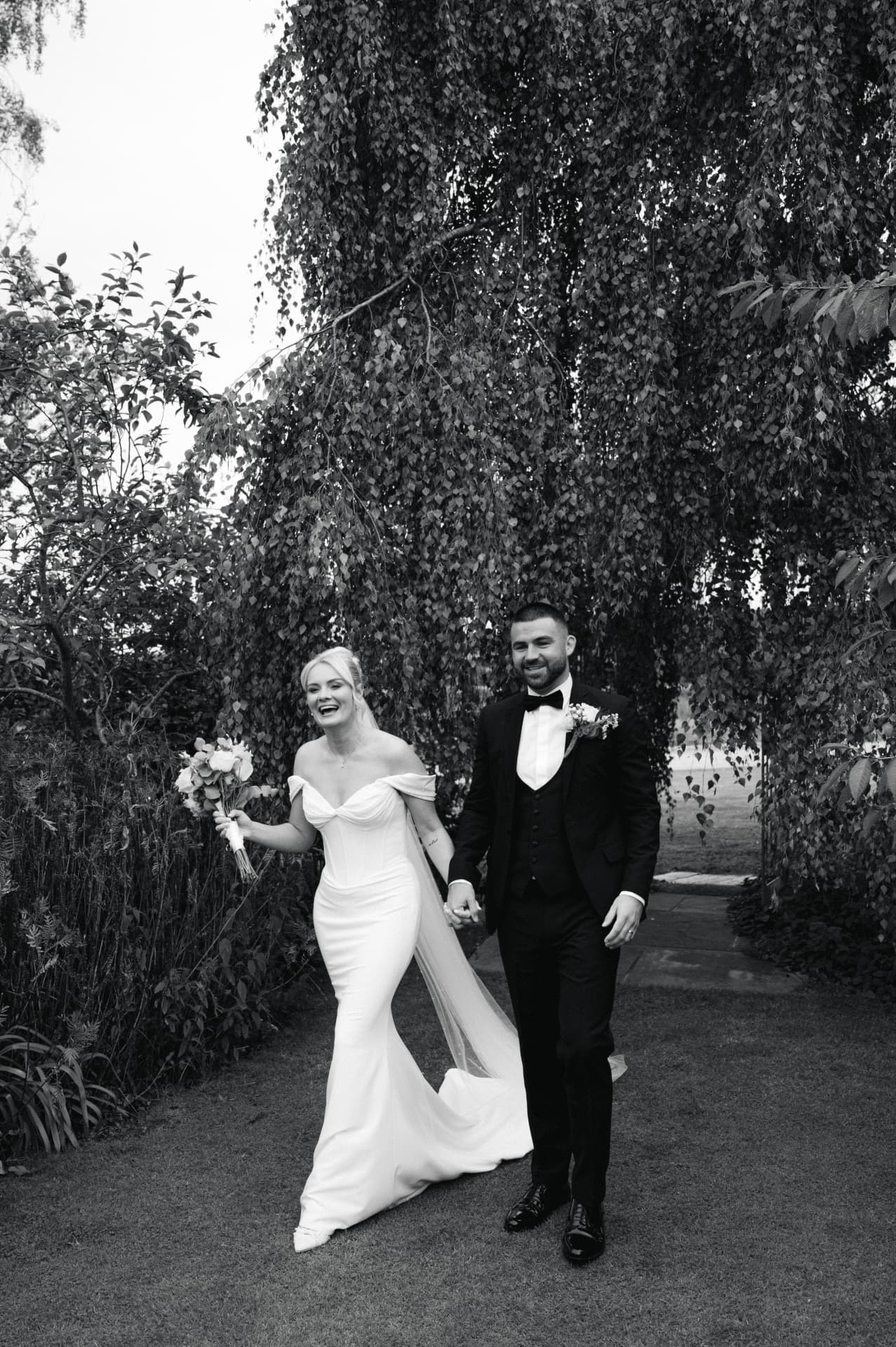 Black and white photograph of a bride and groom laughing as they walk beneath a weeping tree