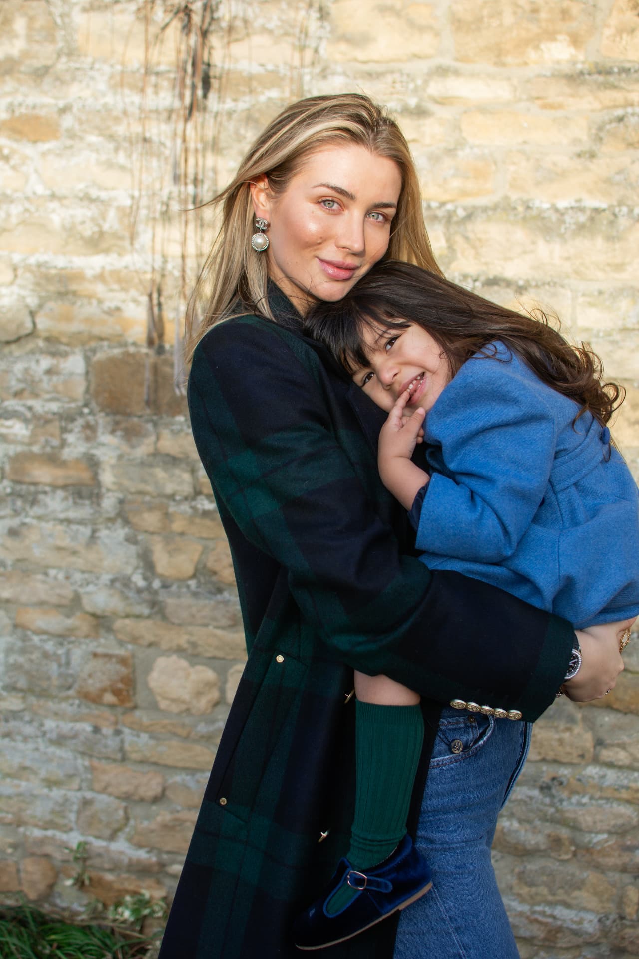 Mother in a plaid coat smiling with her young girl beside a stone wall