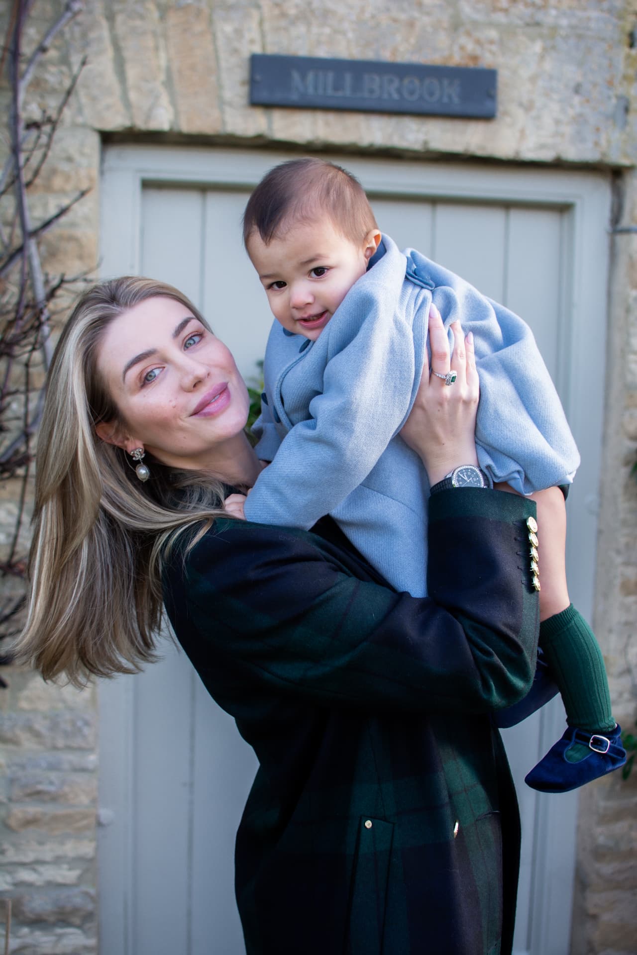 Mother in a plaid coat holding up a young child outside a stone country house