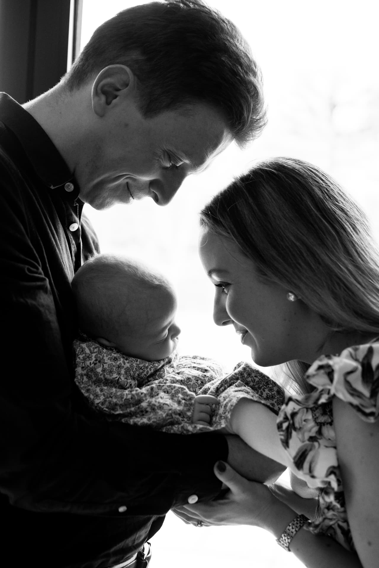 Black and white portrait of parents in profile gazing at their baby by a bright window