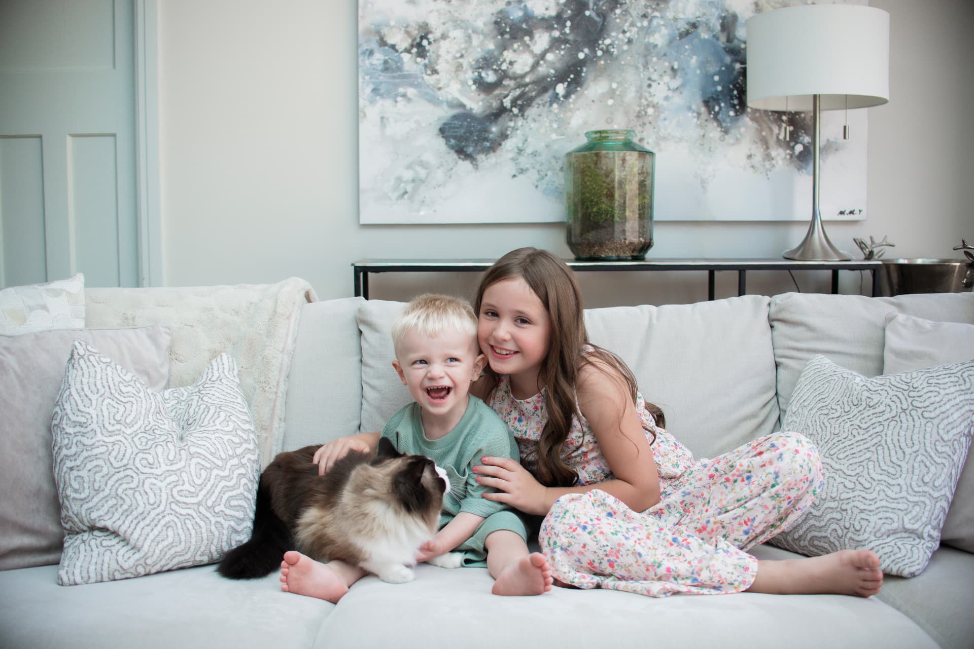 Two children laughing with their cat on a light sofa at home